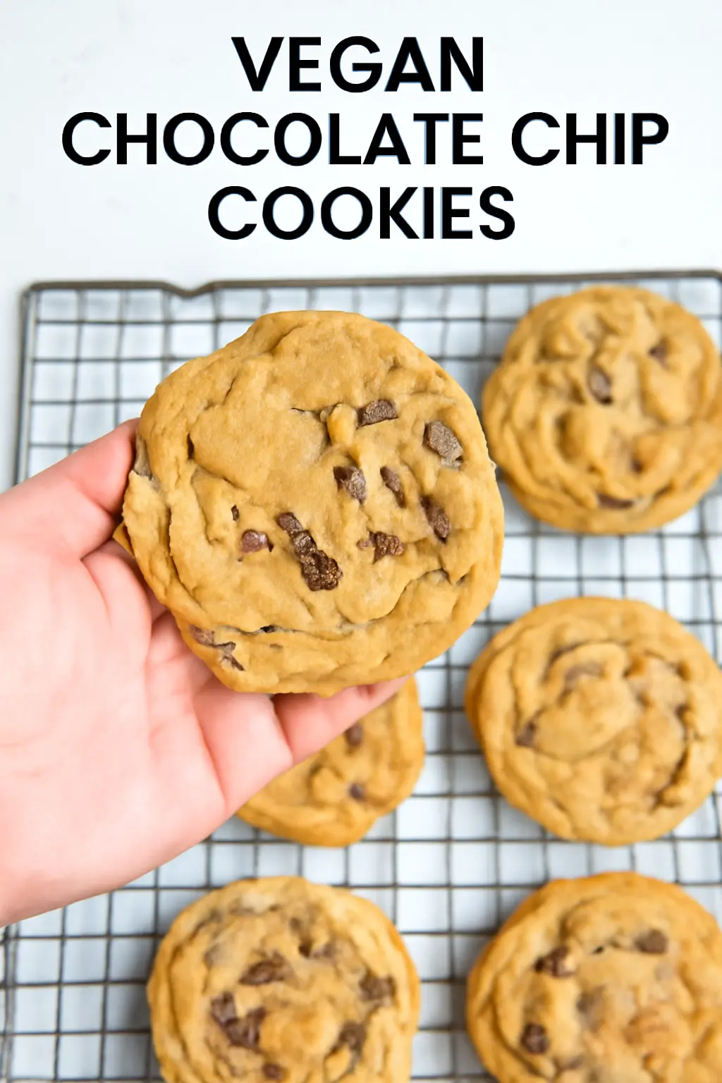 A hand holding a freshly baked Crumbl Vegan Cookies with a soft and chewy texture, showing melted dairy-free chocolate chips. The cookie is placed on a wire cooling rack, highlighting its vegan ingredients and indulgent appeal.
