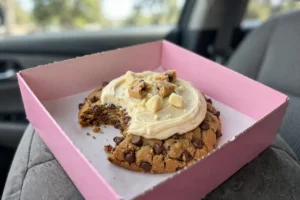 Crumbl Cookie Dough Cake Cookie with a bite taken, in a pink Crumbl box on a car seat
