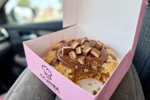 Crumbl Caramel Shortbread Cookie ft. TWIX with a bite taken, in a pink Crumbl box on a car seat